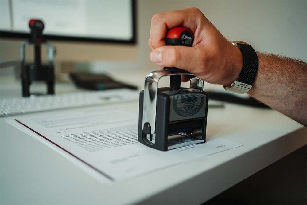 Ein fertiggestelltes Immobiliengutachten wird auf dem Schreibtisch mit einem Stempel abgezeichnet – sinnbildlich für die Qualitätssicherung nach dem Vier-Augen-Prinzip vor der Auslieferung.
